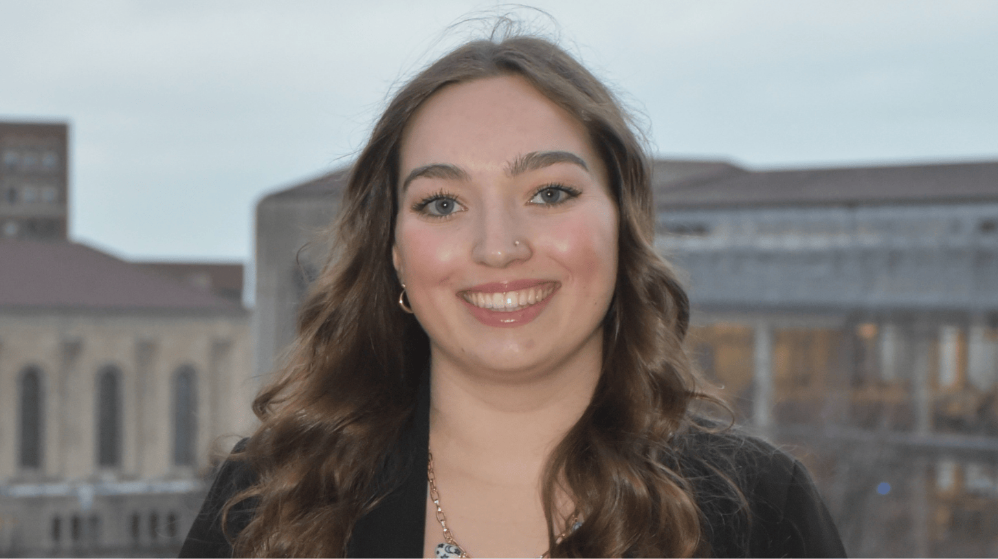 Portrait of a current Gannon Scholar with long wavy brown hair, smiling, wearing a black blazer and necklace, standing outdoors with campus buildings softly blurred in the background.